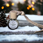 Evergreen Frost Car Diffuser on table with snow and pinecone with Christmas lights in background