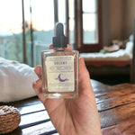 Hand holding Dreamy Bath Body Hair Oil with cozy cabin with cozy beds in background
