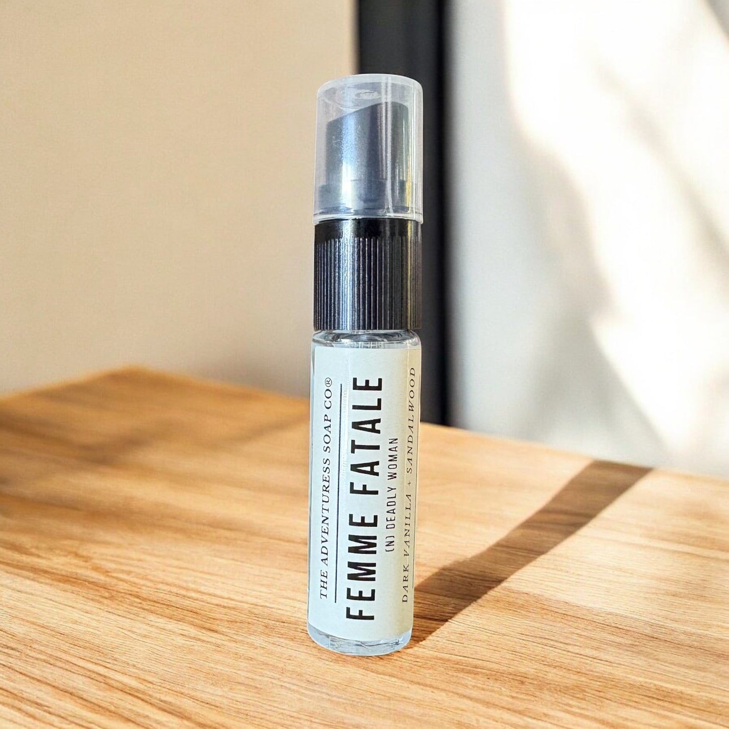 Small spray bottle labeled 'Femme Fatale' on a wooden surface with a neutral background