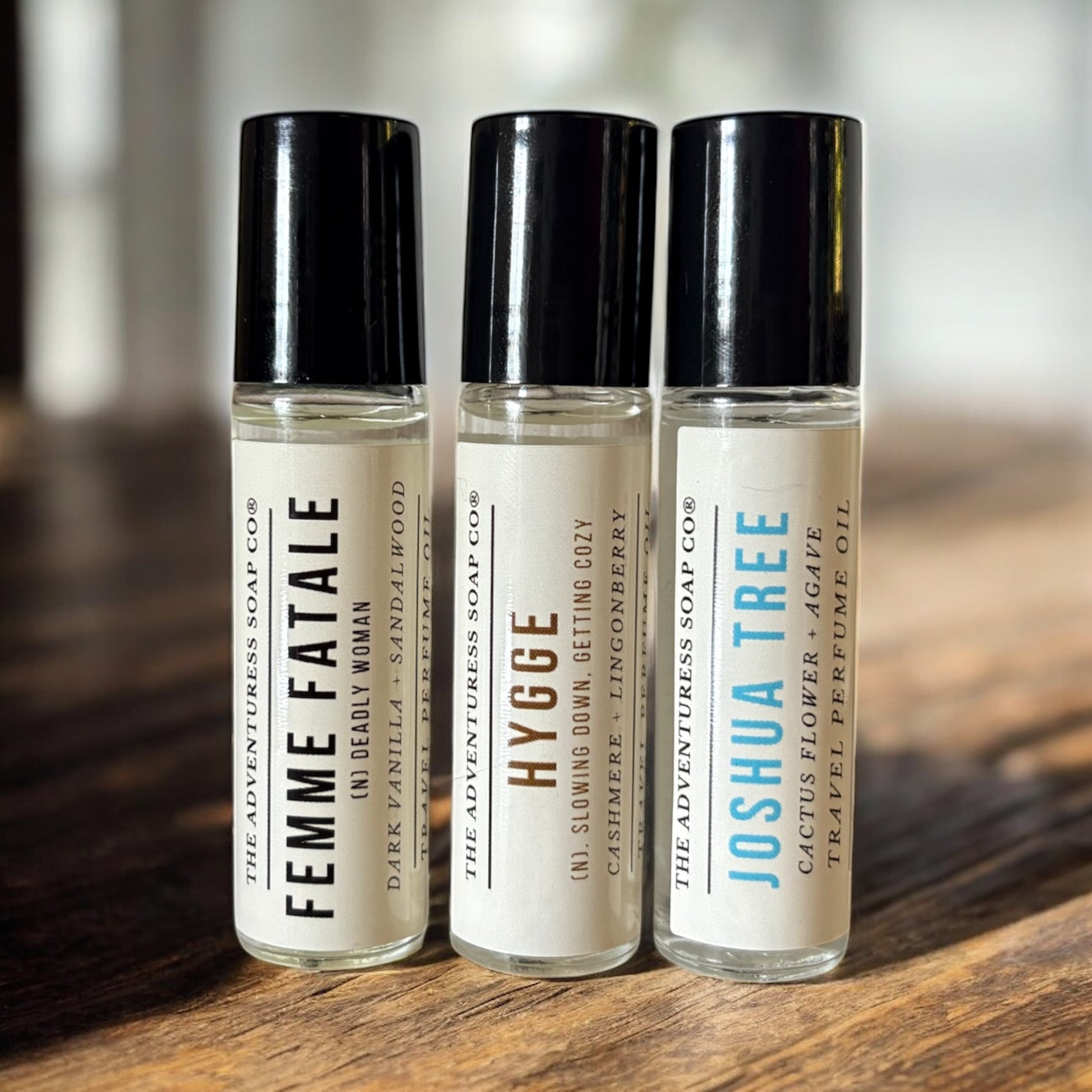 Three perfume travel bottles labeled 'Femme Fatale', 'Hygge', and 'Joshua Tree' on a wooden surface.